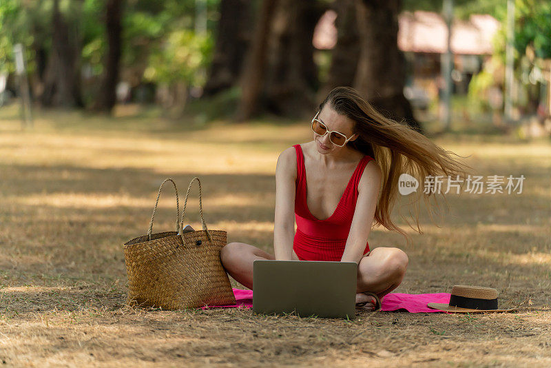 美丽的白人女性性感的女人在红色泳装比基尼戴着太阳镜在她的电脑笔记本电脑上工作，坐在海边的树林里放松