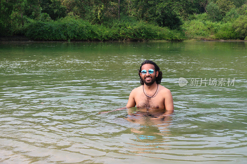 特写照片中，一名印度男子戴着有色的镜面太阳镜，站在齐胸深的泻湖平静的水面上，这是热带岛屿的天堂，可以看到有棕榈树的林地，重点放在前景上