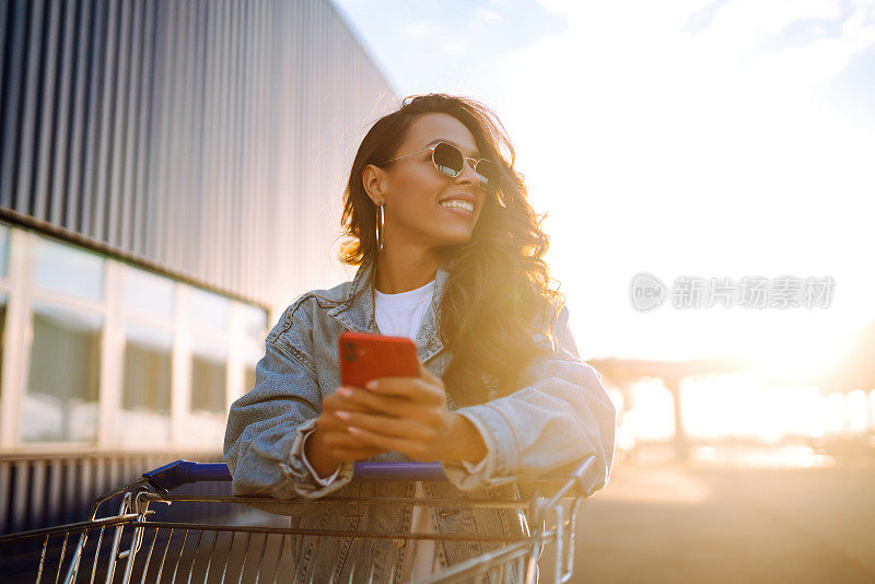 微笑的女人拿着智能手机，推着购物车