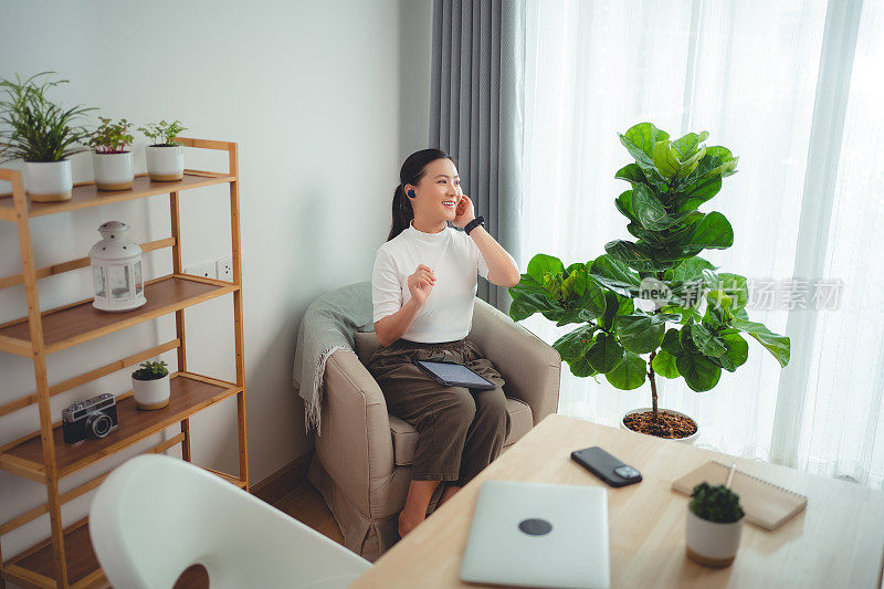 亚洲女性喜欢戴着耳机上网工作，在平板电脑上阅读数据，坐在家里的扶手椅上打电话。