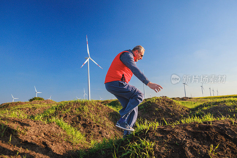 高大的男子在水平轴风力涡轮机旁的绿色山丘上跳跃嬉戏