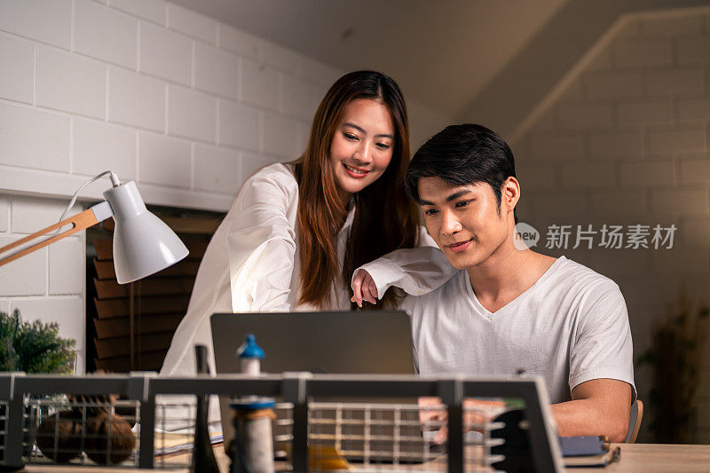 亚洲年轻人晚上在家和妻子在卧室里加班。迷人美丽的妻子穿着睡衣等着丈夫用笔记本电脑工作，在家里睡觉时上网交流。