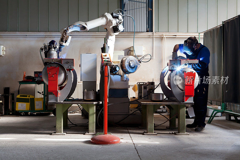 人与机器人在工业建筑中共同工作。机械臂在一名工人的协助下对金属部件进行焊接，该工人反过来手动管理焊接。