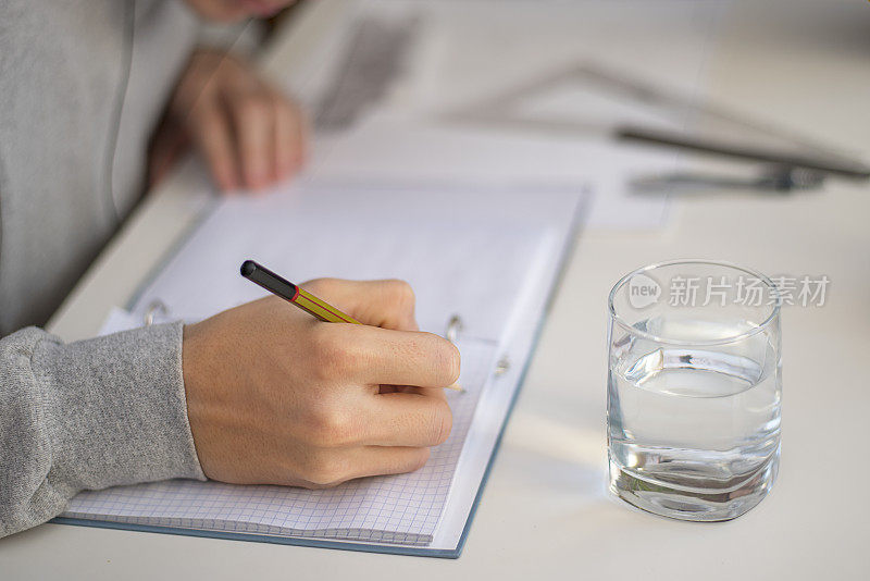 Unknown坐在一张白色的桌子旁，拿着一杯水，用铅笔在记事本上写字