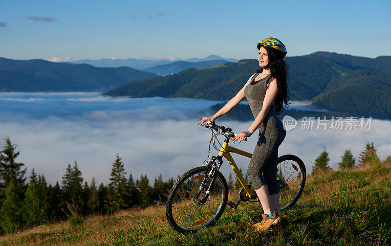 迷人的女子骑着黄色的自行车在山上骑行，戴着头盔，享受着夏日清晨的日出。雾蒙蒙的山，模糊的背景上的森林。户外活动。本空间