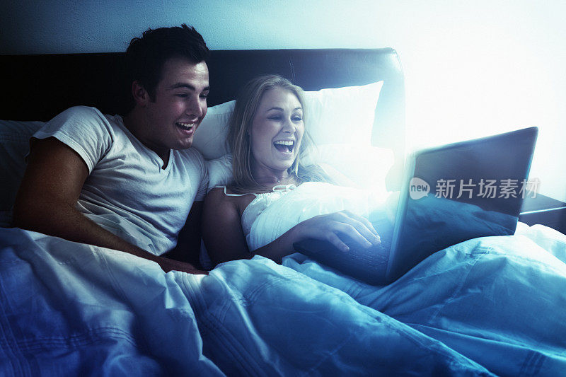 快乐的年轻夫妇坐在床上看笔记本电脑和大笑