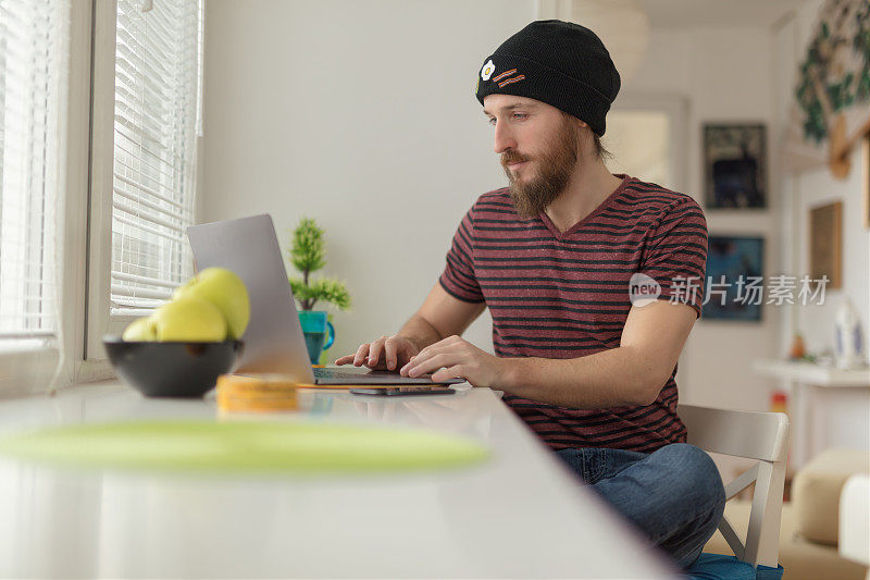 一个留着胡子戴着冬帽的年轻人在靠窗的客厅里用笔记本电脑工作，喝着咖啡。