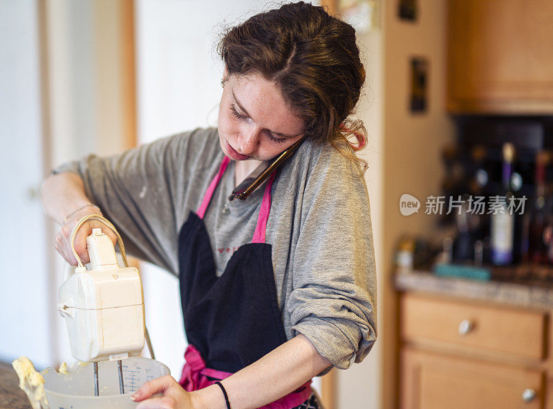 一名年轻女子正在自家厨房里烤香蕉面包，她一边用电动搅拌器搅拌各种配料，一边还在讲电话。