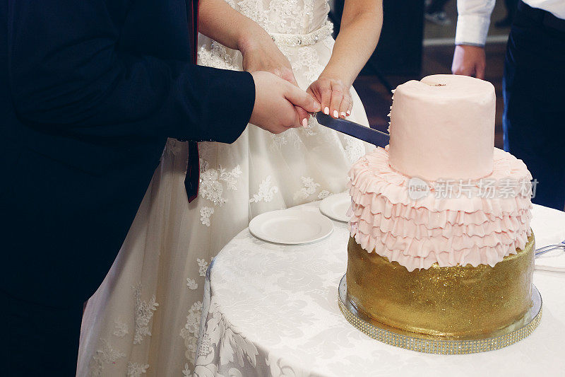 快乐的新婚夫妇在餐厅的婚宴上切着镶有金色和粉色糖霜的美味婚礼蛋糕，快乐的新娘新郎时刻