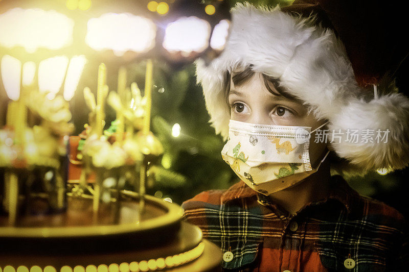 小男孩戴着圣诞老人帽和防护面具看着一个玩具旋转木马