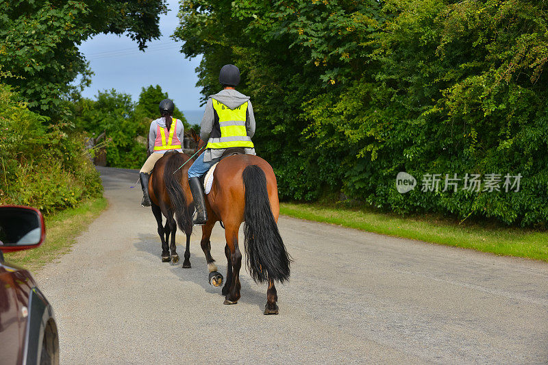 一对年轻的夫妇正享受着骑马的乐趣，他们穿着亮黄色的高维安全装置在威尔士乡村的乡间小路上骑行，这样他们就能被其他道路使用者看到，比如跟在他们后面的汽车。