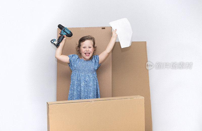 一个6岁的白人女孩站在大纸箱的背景下，手里拿着一个无绳螺丝刀。