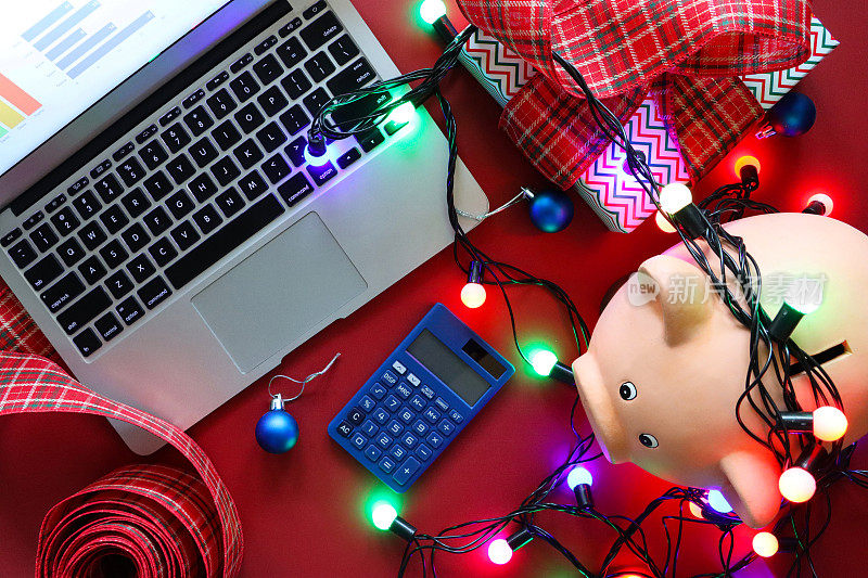 形象的小猪银行与照明线仙女灯在红色表面，笔记本电脑和蓝色计算器包装圣诞礼物与格子丝带蝴蝶结，升高的观点，圣诞预算，账单，债务和财务概念