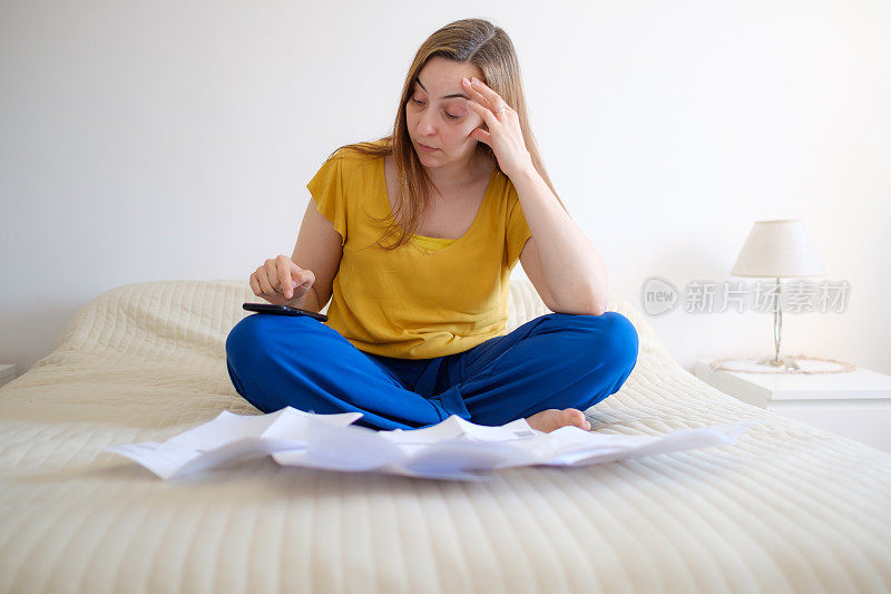 女人检查她的账单她的账单在她的床上，用手机计算器做数学，变得担心