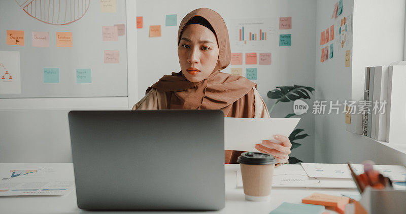 年轻的亚洲穆斯林女商人，戴着头巾，坐在办公室的办公桌前用笔记本电脑工作。