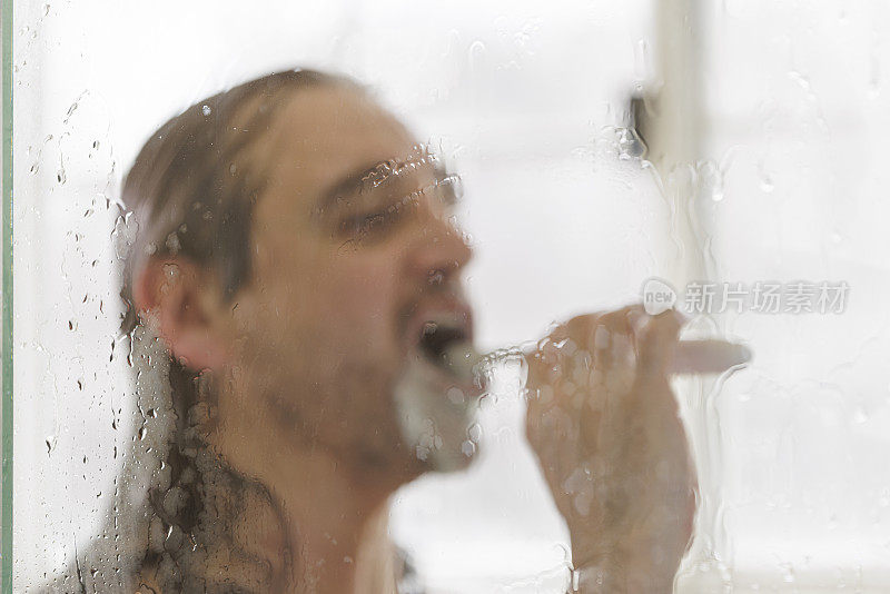 成熟的50岁快乐男人在洗澡时刷牙。脸是散焦的，焦点在淋浴门的湿玻璃上。