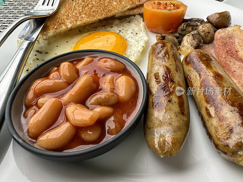 全帧图像的白色盘子包含全英式油炸早餐食材，香肠，烤豆小蛋糕，培根片，番茄，蘑菇，煎蛋，吐司片在餐厅的桌子上，刀和叉，抬高视图，重点在前景