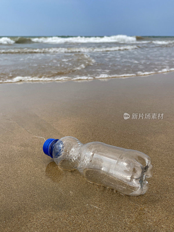 特写图像，空的，透明的塑料饮料瓶，蓝色的盖子被冲到沙滩上，在退潮水的边缘，破碎的海浪冲刷海洋垃圾和岸上的污染，乱扔的沙子，肮脏的海滩，关注前景，复制空间
