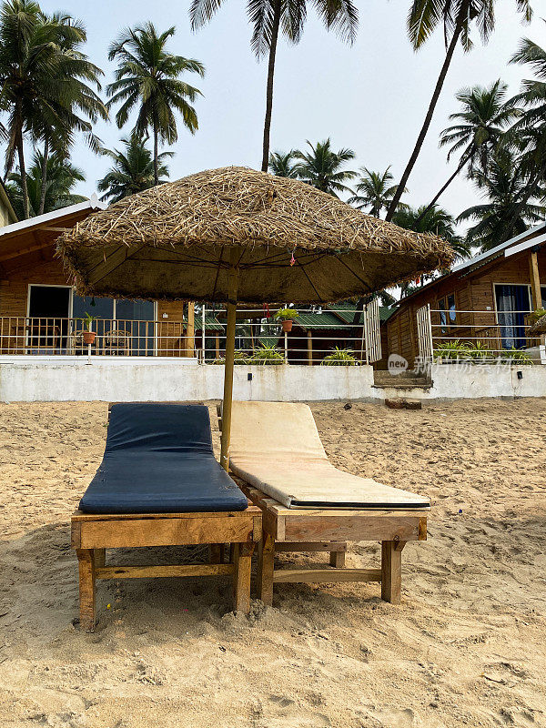 海滩度假的图像，一对木制的，蓝色和奶油色软垫的太阳躺椅床，在稻草遮阳伞下在沙滩上晒太阳，在蒸汽船上擦拭干净的垫子，一排海滩小屋，果阿，印度