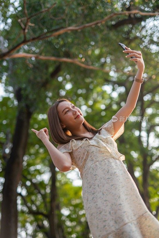 年轻的亚洲女子在公园用手机自拍。在郁郁葱葱的绿色森林里，美女视频通话