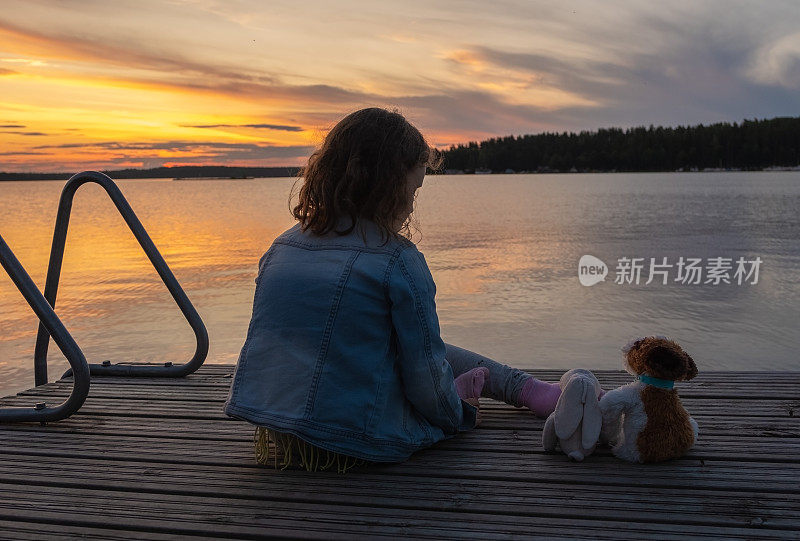 8岁的小女孩拿着毛绒玩具坐在湖边的木码头上看日落。是时候做梦了。