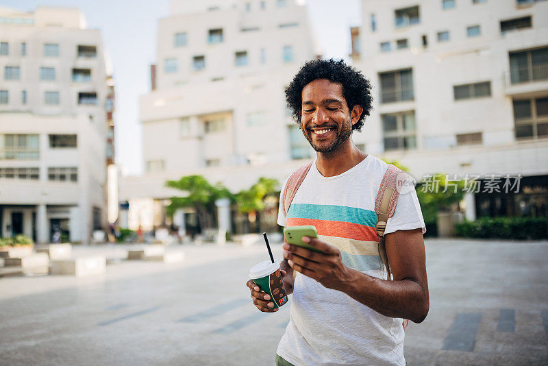 一位面带微笑的男游客在城市里走来走去，一边用着手机