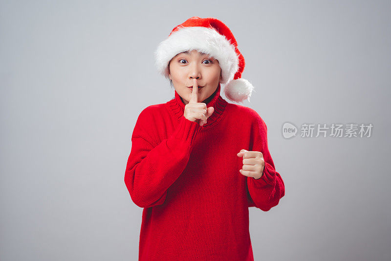 亚洲女人戴着圣诞老人的帽子，开心地微笑着，把食指放在嘴唇上，孤立地站在白色的背景上。