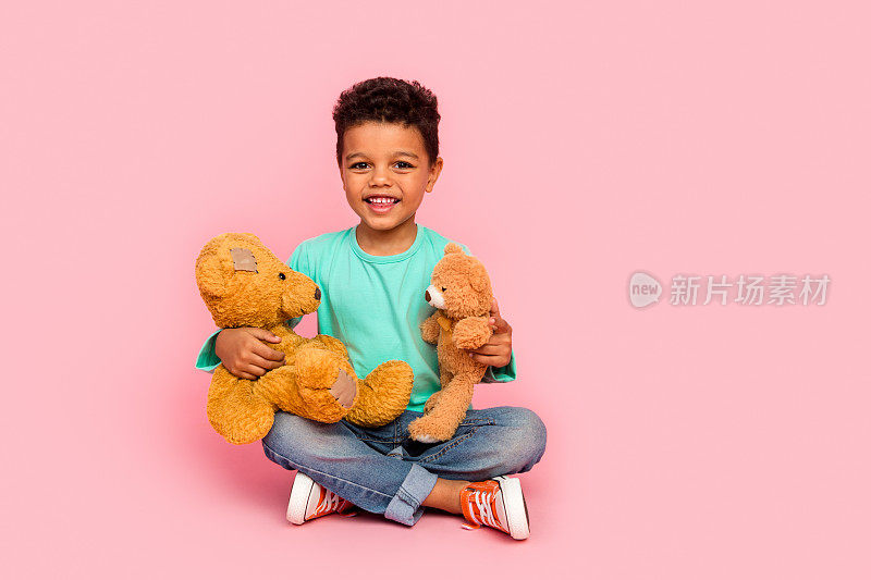 可爱小男孩的全身照片坐在地板上玩两个泰迪熊穿着时髦的海蓝宝石衣服孤立在粉红色的背景