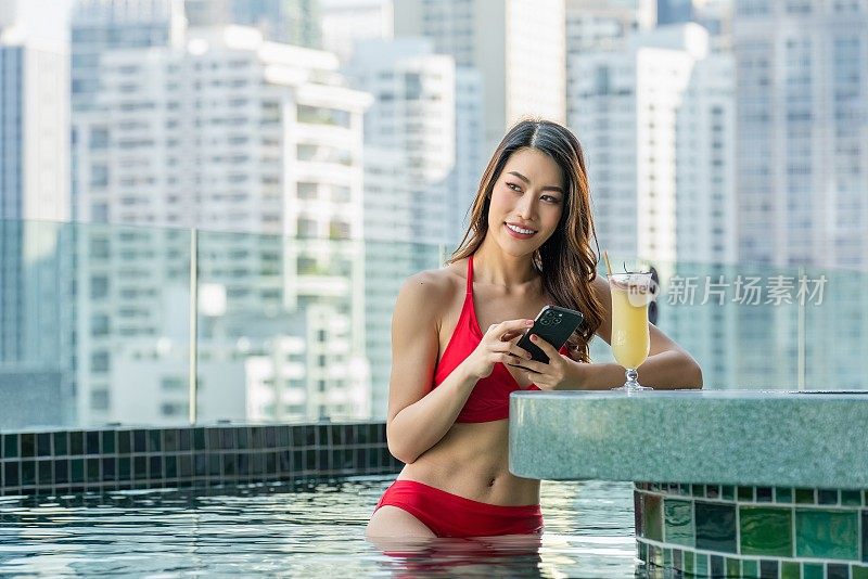 泳池边的乐趣:年轻女子坐在屋顶泳池酒吧与城市景观的背景，享受饮料和使用她的手机。