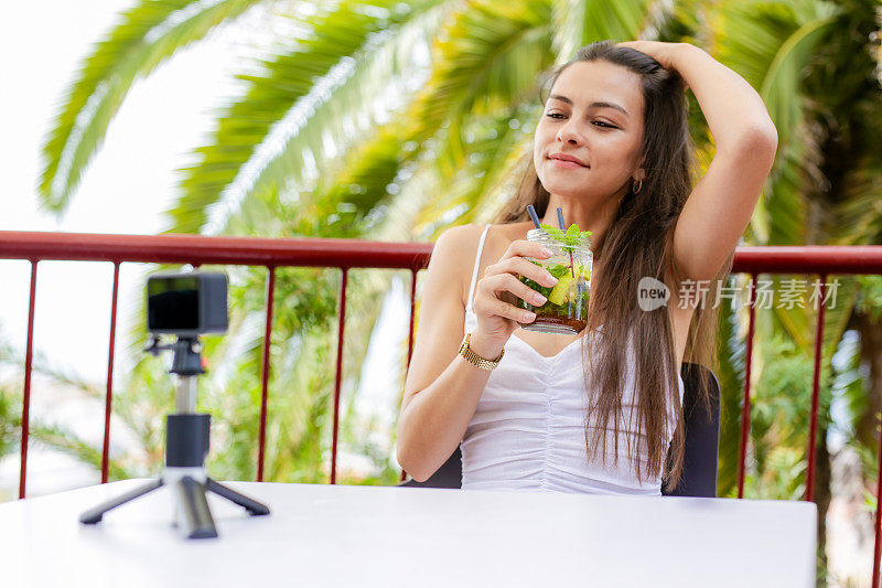 年轻女子坐在面向大海的露台上记录博客。在现代咖啡馆拍摄社交媒体博客