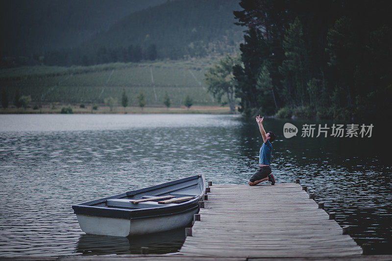 一个男人跪在一个美丽的湖上的木制码头上祈祷投降，在大自然中有一艘系泊的划艇