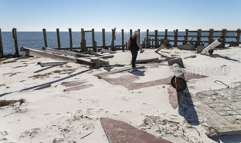 一个成熟的白人女人在海滩上走在被飓风摧毁的房子上。