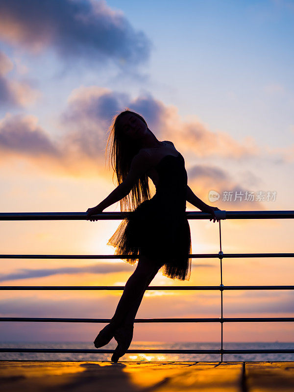 在日出或日落时，穿着黑色芭蕾舞裙的芭蕾舞女演员的剪影在海洋或海上的堤岸上。年轻迷人的金发女人长头发练习伸展和练习
