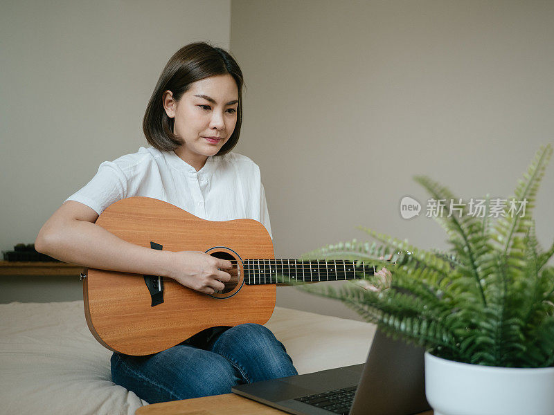 年轻女子微笑着坐在床上拿着吉他看着笔记本电脑