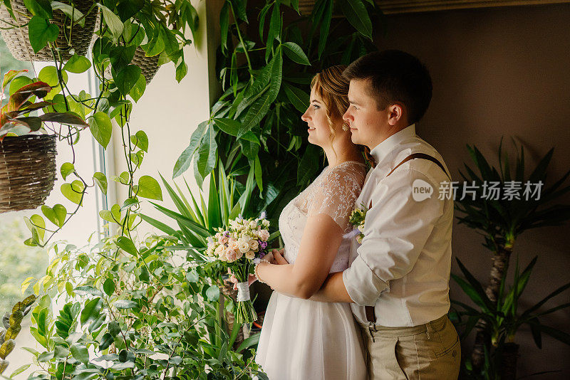 新婚夫妇相依相拥，望着窗外，正对着室内的绿植。