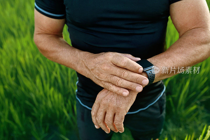 运动男子在户外跑步后检查智能手表。在稻田里的草地上，用手腕上的健身追踪器拉近男性的双手。