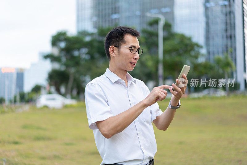 一位亚洲男性白领正在办公大楼外的草地上使用智能手机