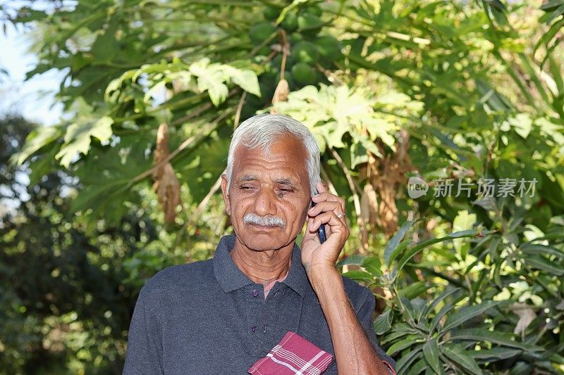 一位印度资深农民站在花园或田地里，用付费手机听取呼叫中心农业官员的建议