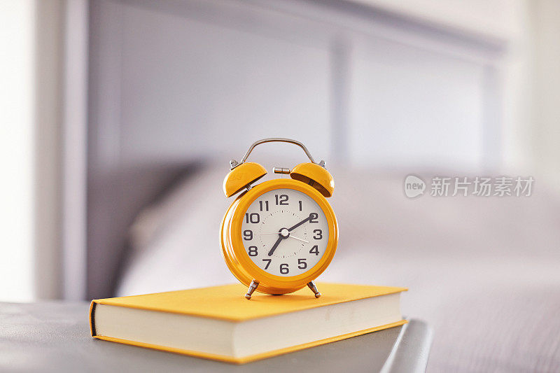 在现代明亮的卧室里，明亮的黄色闹钟和书放在床头柜上