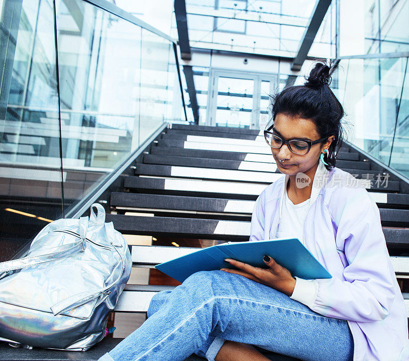 年轻可爱的印度女孩在大学大楼的楼梯上看书，戴着时髦的眼镜，生活方式的人的概念近距离