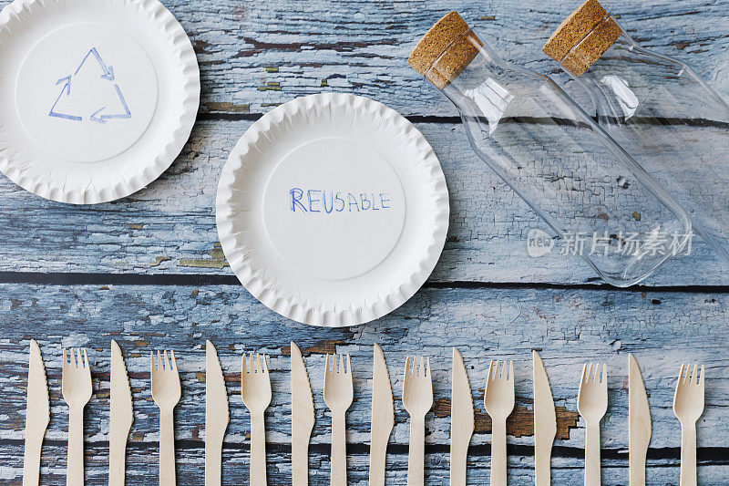 浅蓝色木制背景上的环保餐具平面组合。牛皮纸盘子和木制餐具。可回收的纸制品，零浪费的理念。有选择性的重点