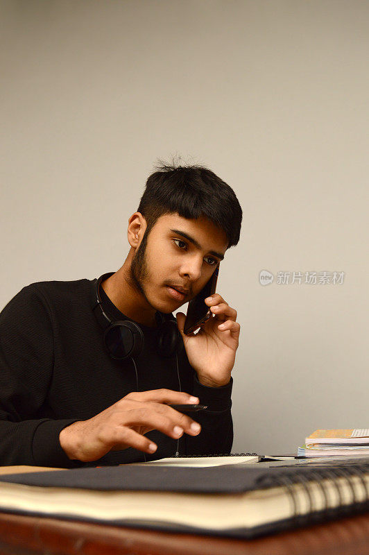 一个年轻聪明的印度亚裔青少年男孩的垂直镜头集中注意力，而接听或交谈的手机拿在他的手在灰色的背景上，并做笔记，同时写在笔记本上