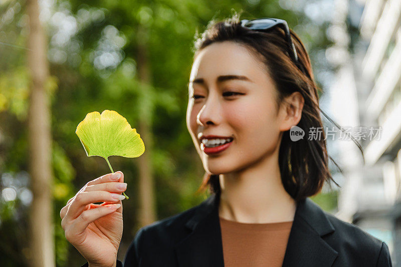 一个迷人的女人站在城市的街道上，手里拿着一片银杏叶。