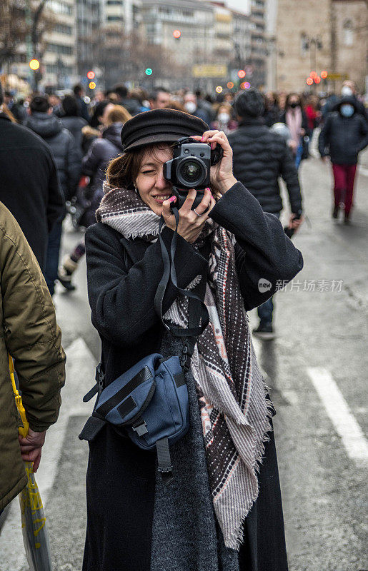女孩穿着黑色外套和贝雷帽，在拥挤的街道上用单反相机拍照。版本