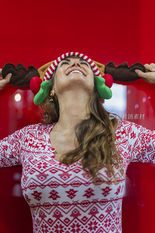 平均年龄40岁的拉丁裔妇女站在红色背景墙上，穿着圣诞睡衣和驯鹿帽