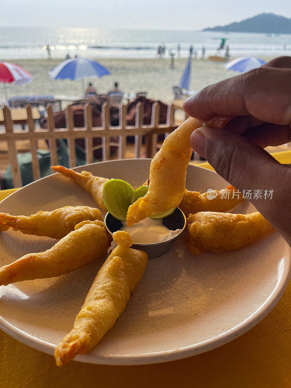 特写图像无法识别的人蘸天妇罗虾在碗蛋黄酱，露天用餐场景，炸虾与楔形石灰，海滩海鲜餐厅，海边日光浴躺椅，海滩阳伞背景，重点在前景