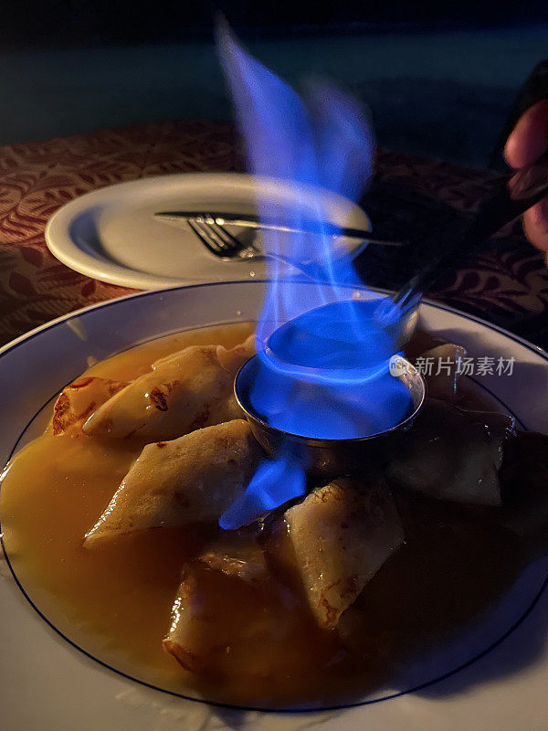 特写照片是一盘法国薄饼（薄煎饼），上面覆盖着橙色和焦糖酱，奶油酥饼配橙色利口酒，蓝色火焰放在金属酒精碗里，聚焦前景