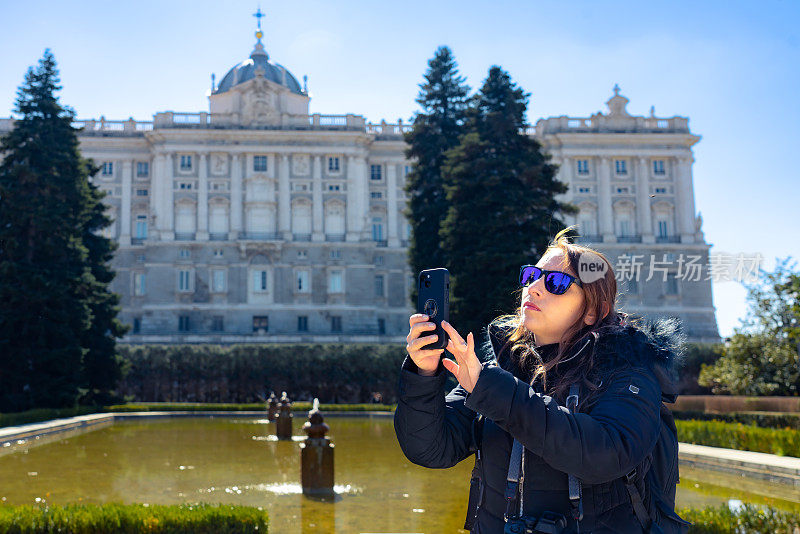 西班牙马德里，年轻的西班牙女游客穿着保暖的衣服，戴着墨镜，坐在绿树成荫的花园里，拿着智能手机和专业相机，对着王宫拍照