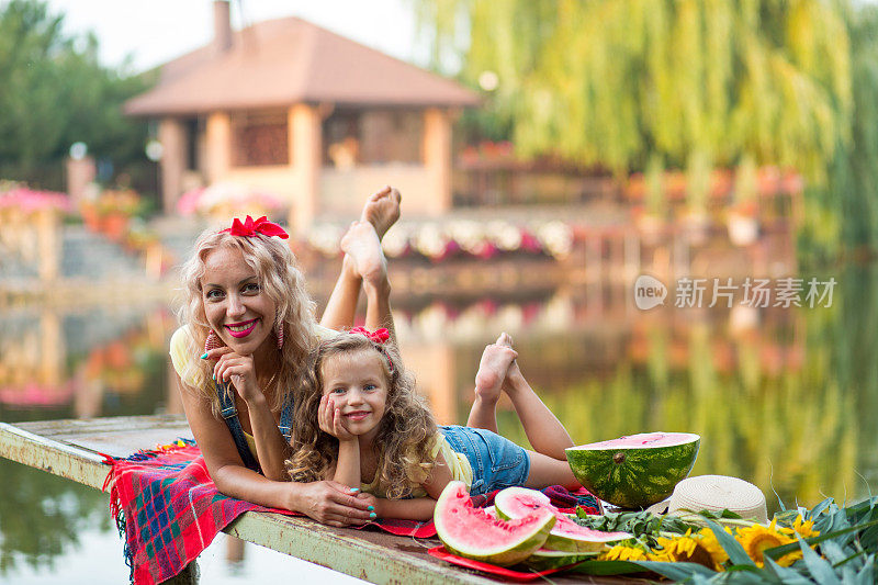 金发碧眼的母亲和三岁的女儿穿着牛仔短裤，带着红色的蝴蝶结，坐在码头上拥抱在一起，码头上有一个切好的西瓜和一束向日葵，背景是河流