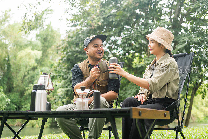年轻的亚洲夫妇，男人和女人坐在户外露营地喝咖啡。两个人带着咖啡机工具在森林里露营。旅游，放松，露营，度假，假日，周末的主题